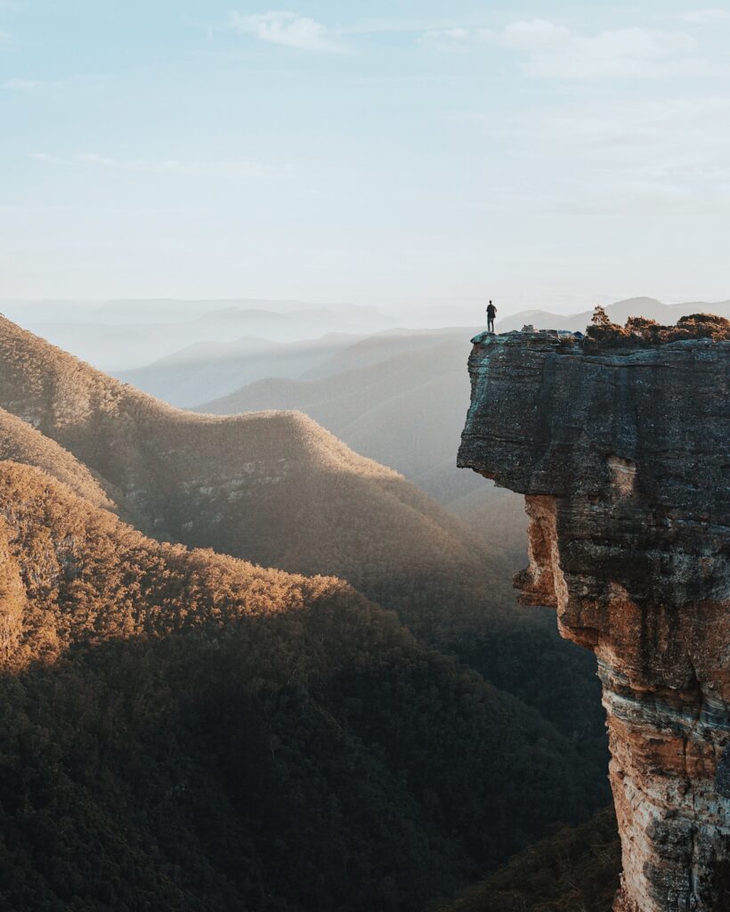Adventurer standing alone on a rocky clifftop overlooking expansive forested valleys and mountains.
