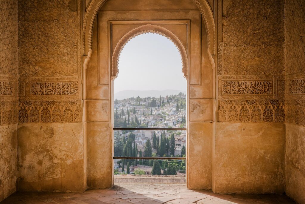 View through an ornate arched doorway of a sun-lit Spanish village nestled on a hillside below.