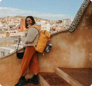 Woman with orange backpack overlooking a Spanish hillside town from a terracotta rooftop.