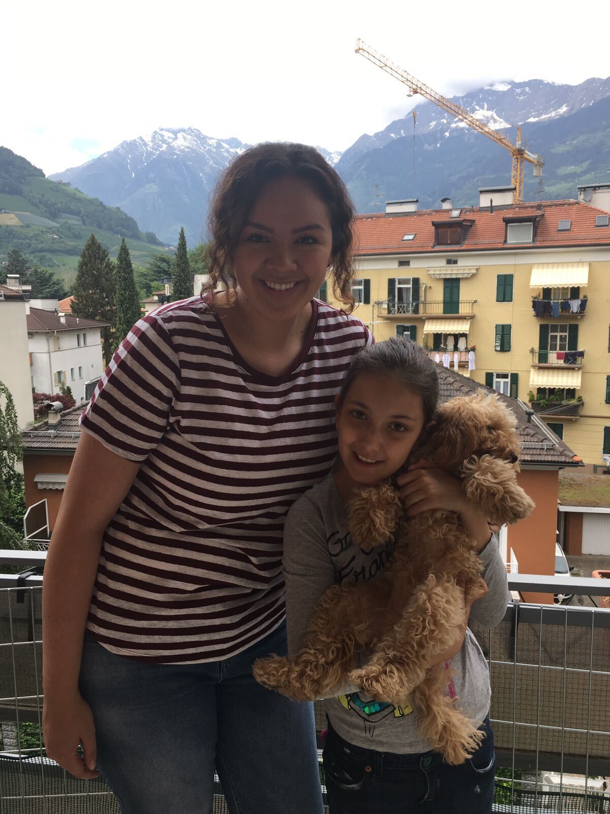 Smiling young woman with child holding a small brown dog on an Alpine balcony.