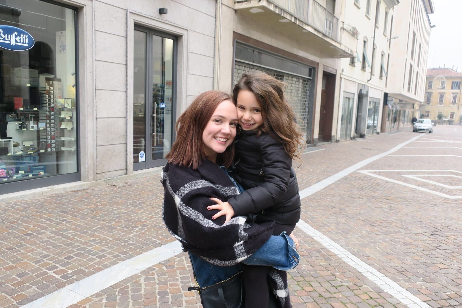 A young woman happily holds a child on a sunny Italian cobblestone street.