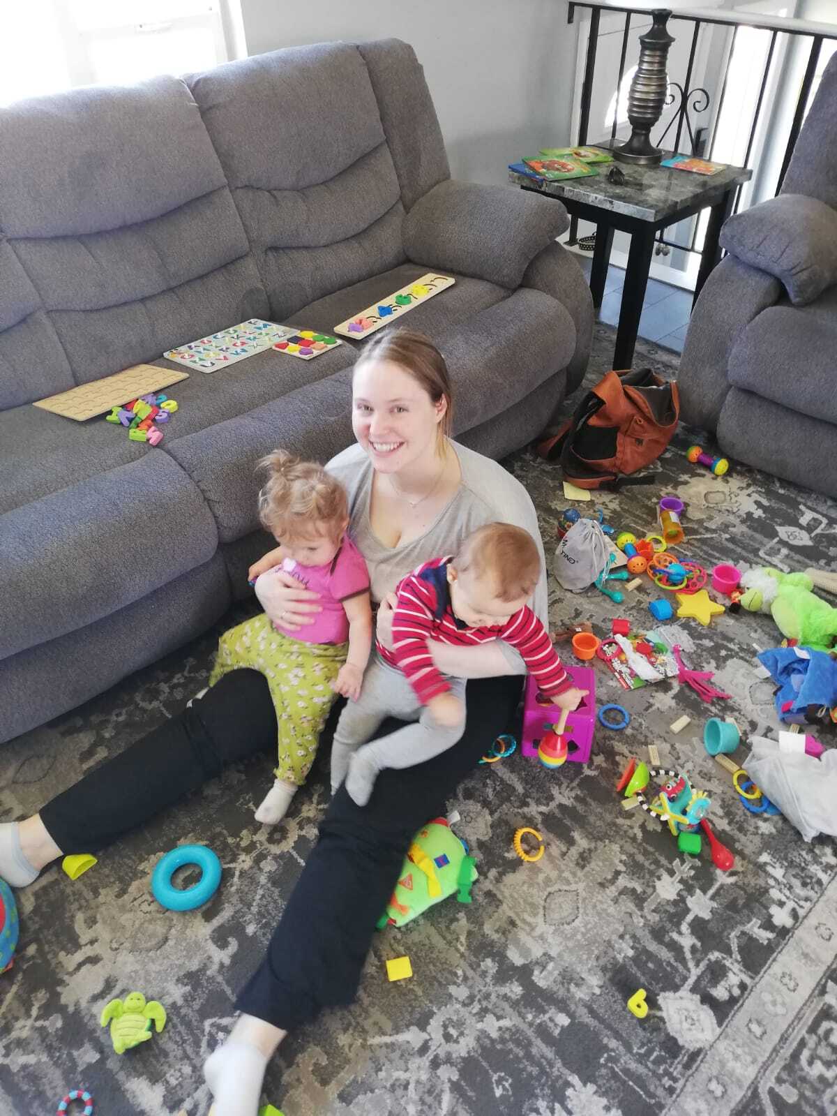 Au pair playing with two toddlers among colorful toys in a bright living room.