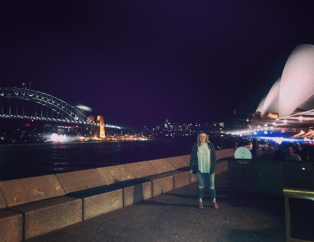 Young woman smiling by Sydney Harbour at night with the illuminated Opera House and Harbour Bridge.
