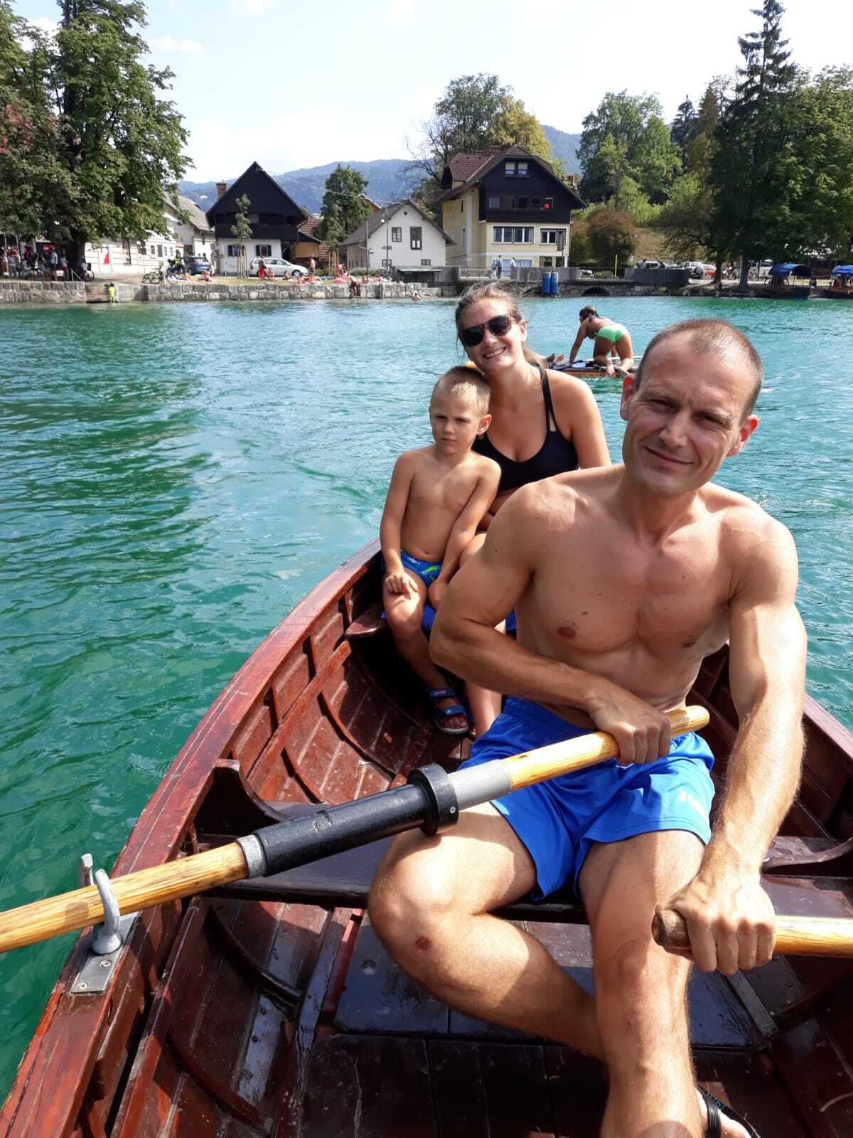 A man rows a wooden boat with a woman and child on a turquoise Alpine lake surrounded by mountains and chalets.