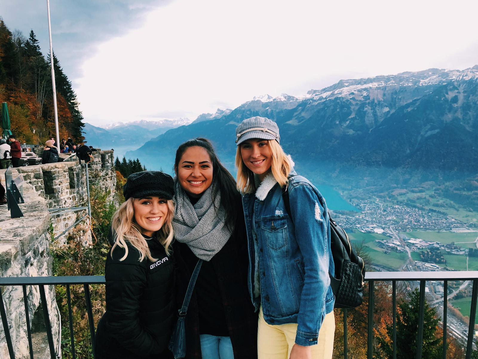 Three young women smiling at a scenic mountain overlook with a turquoise alpine lake and snow-capped peaks below.