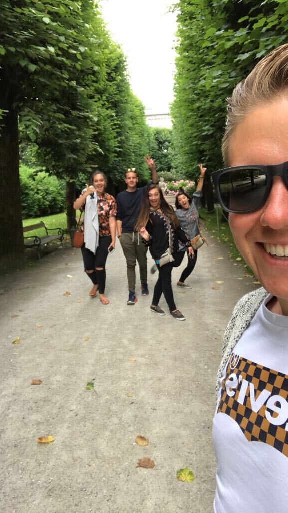 Young woman taking a selfie with friends along a tree-lined European avenue.