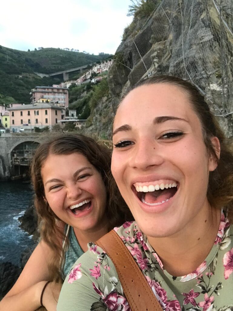 Two young women laugh together in a selfie with a colorful hillside Italian village and river behind them.