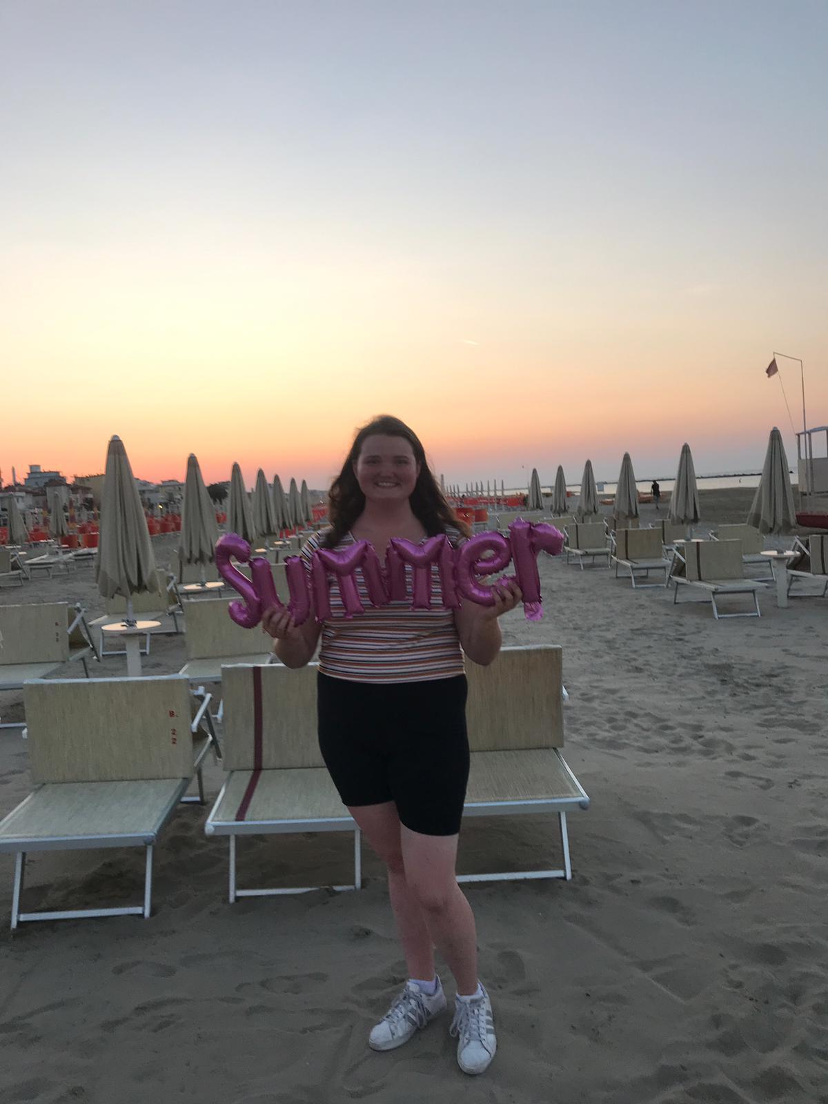 Smiling woman holding purple balloons on an Italian beach at sunset.