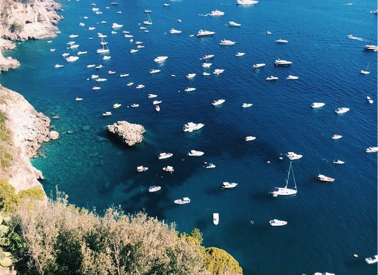 White sailboats anchored in crystal-clear turquoise Mediterranean waters beneath white cliffs.