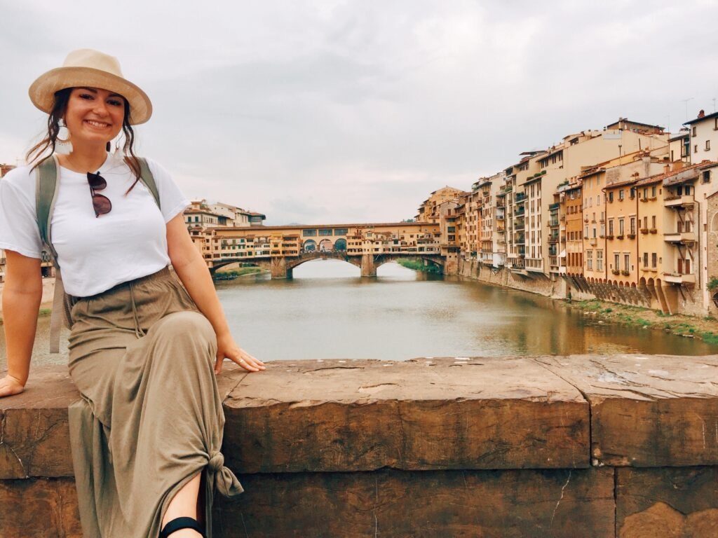 Young woman smiling by the Arno River in Florence with the Ponte Vecchio bridge behind her.