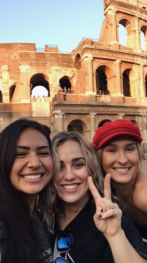 Three young women smiling together at an ancient Italian amphitheater with honey-colored stone arches.