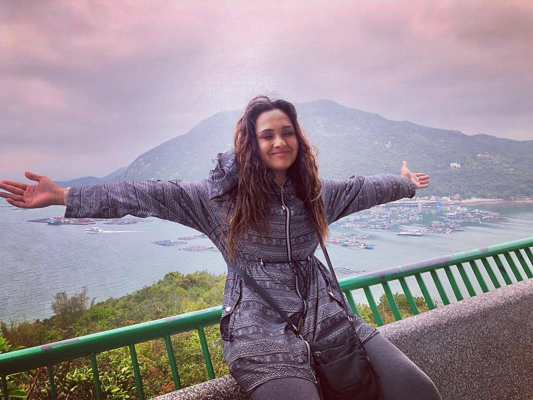 Young woman with arms spread wide at a coastal mountain viewpoint overlooking a bay with fishing boats.