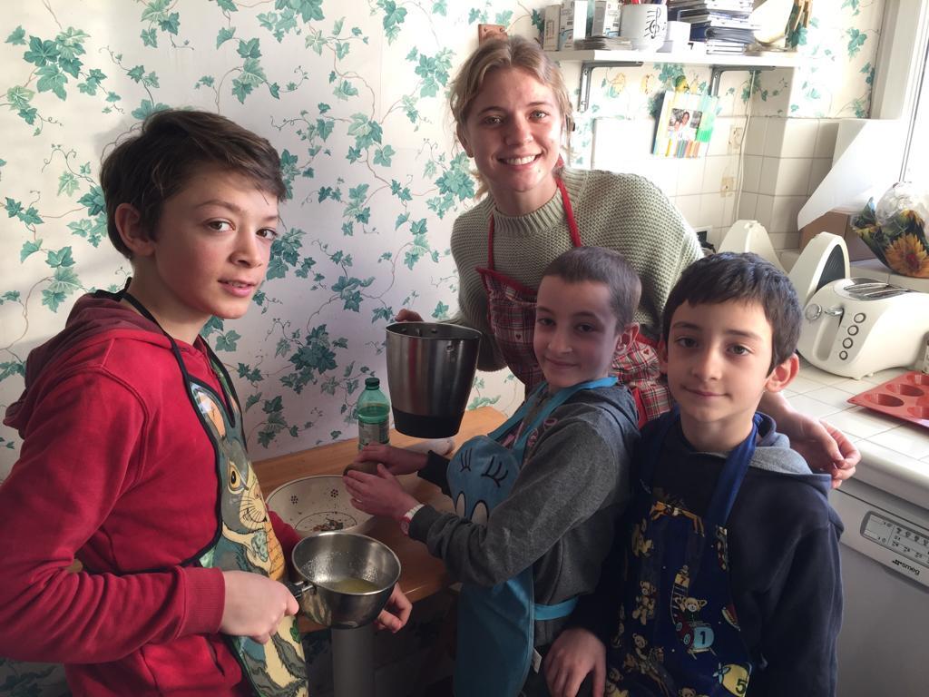 Woman smiles while cooking with three children in a bright kitchen with plants.