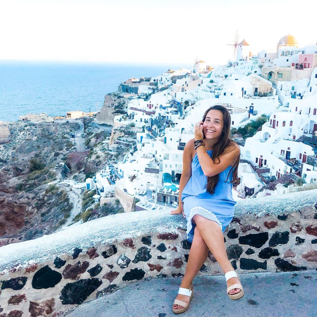 Woman smiling on clifftop overlooking whitewashed Mediterranean coastal village.
