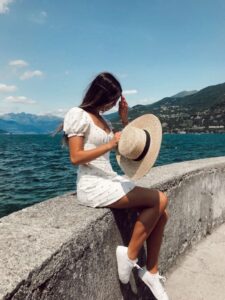 Woman in white dress and straw hat overlooking a turquoise Alpine lake with mountains.