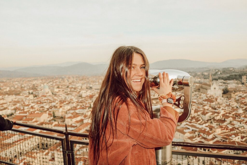 Young woman smiling while gazing through a telescope overlooking a sprawling Mediterranean cityscape.