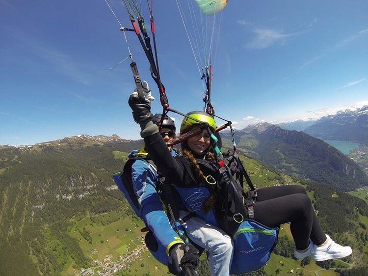 Two young women paragliding over alpine mountains with verdant valleys and a lake visible below.