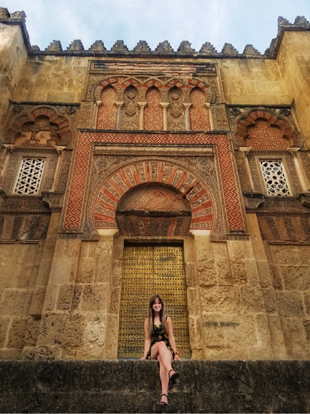 Young woman smiling before an ornate historic building with Islamic geometric tilework and brass door.