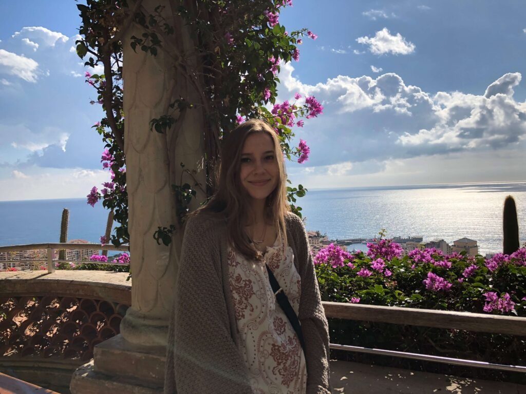 Young woman smiling beside magenta bougainvillea with Mediterranean seascape behind.