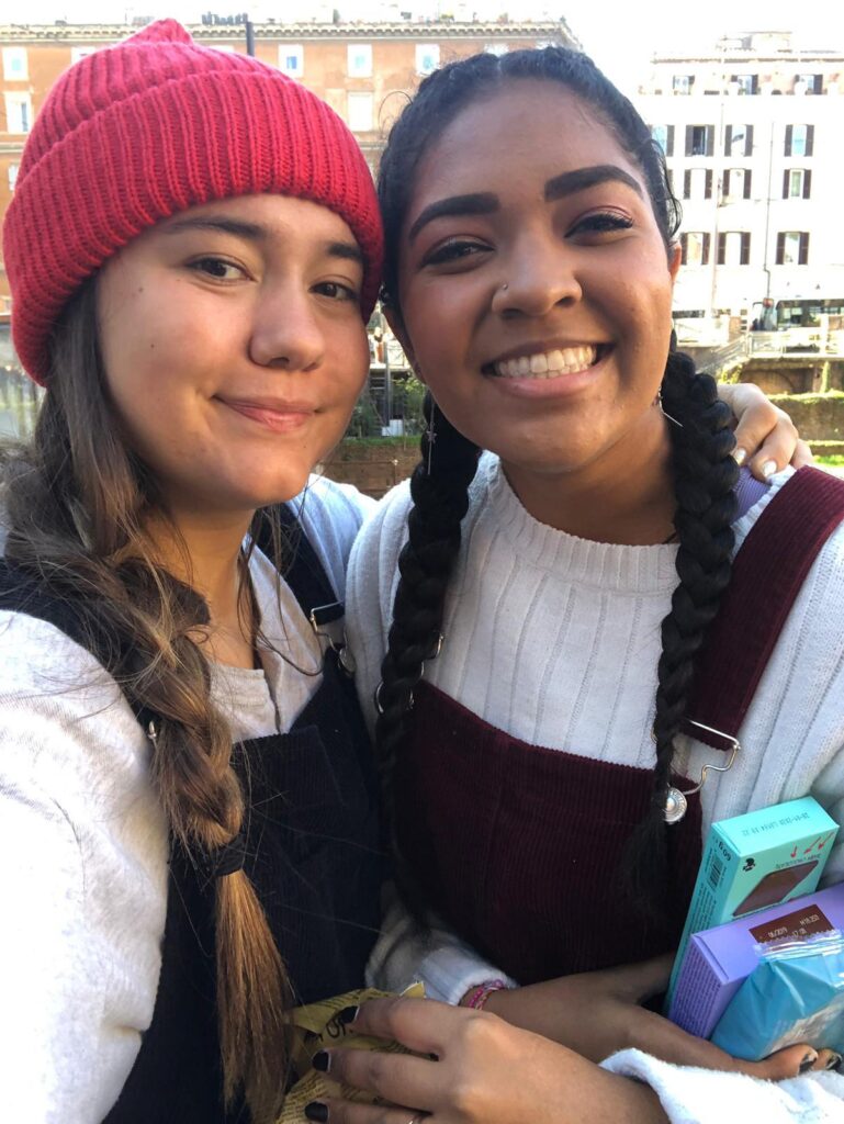 Two young women smile together with historic European city architecture visible behind them.