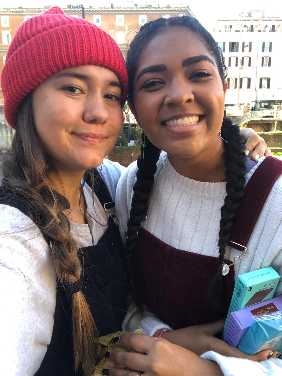 Two young women smile together with historic European city architecture visible behind them.