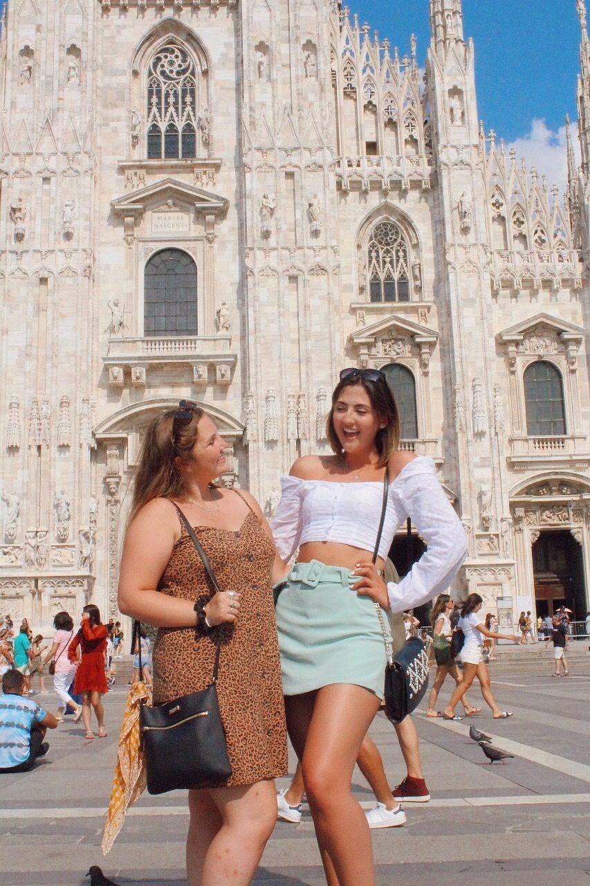 Two young women smiling at each other in front of Milan Cathedral, exploring together in Italy.
