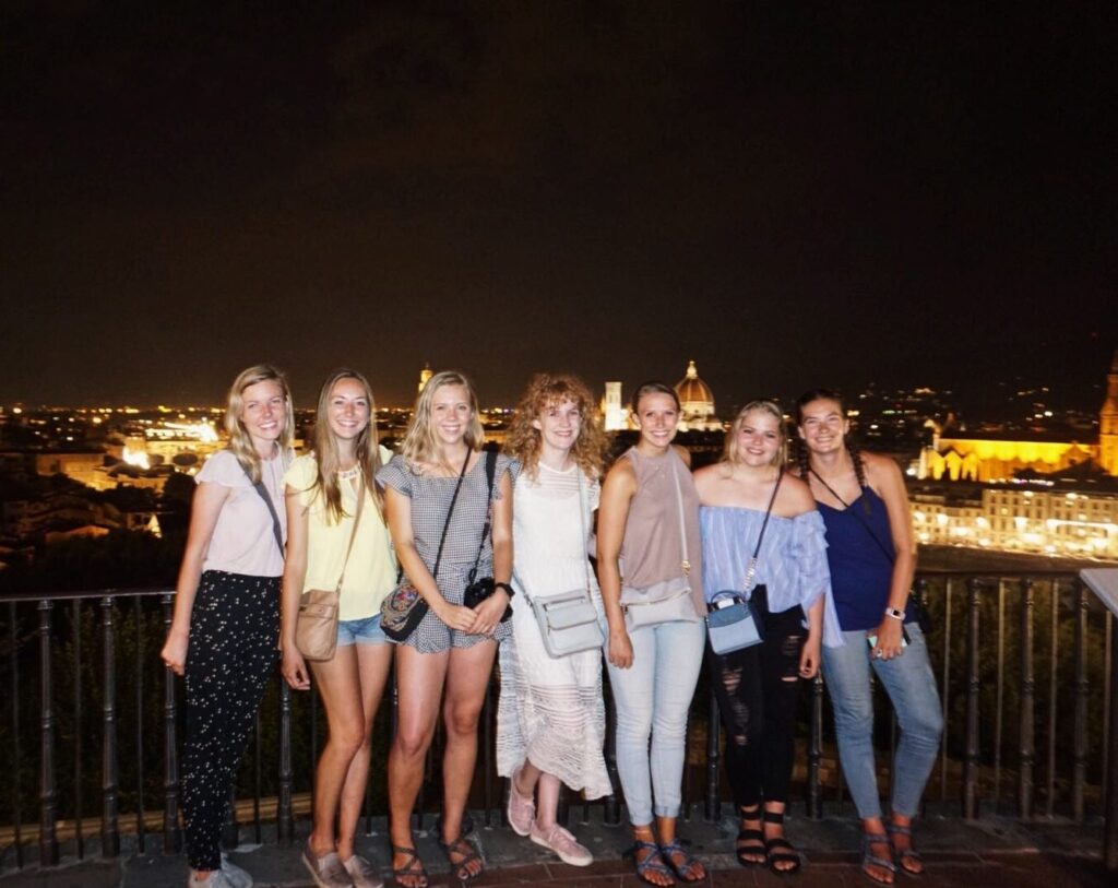 Young women smile together on a Florence rooftop with the illuminated cathedral glowing behind them.