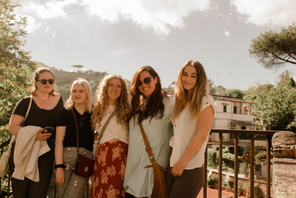 Five young women smiling on an Italian hillside terrace with Mediterranean landscape and traditional buildings.
