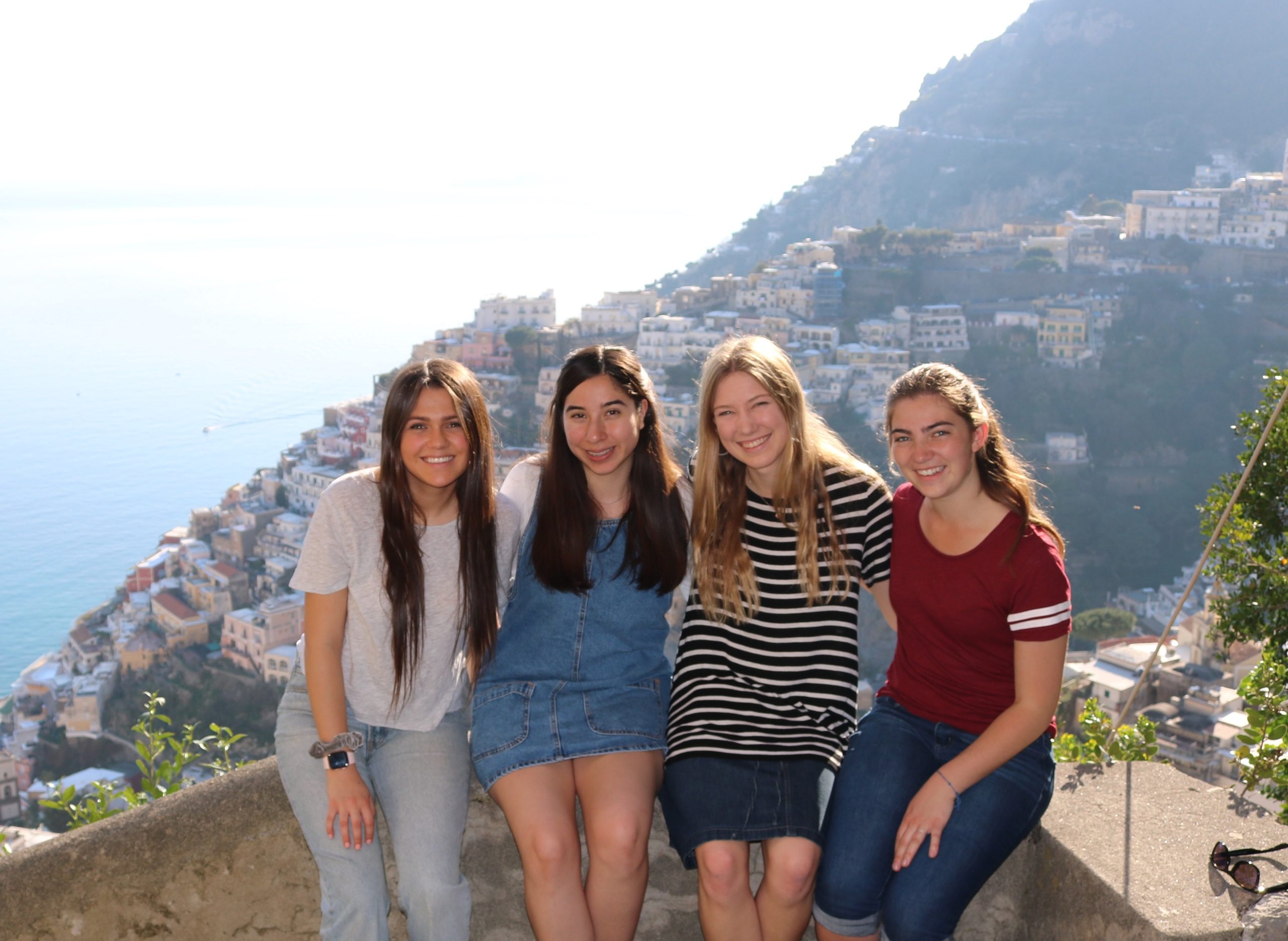 Four young women posing with a Mediterranean coastal town cascading down the hillside.