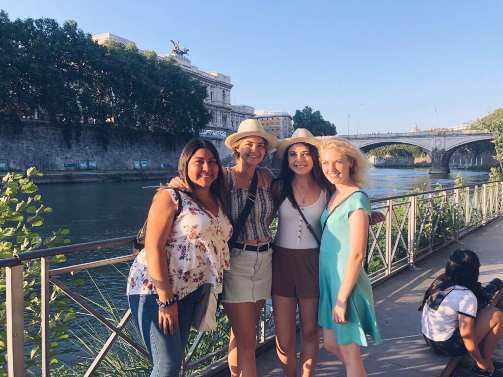 Four young women smiling together on a Rome riverside promenade with historic monuments in the background.
